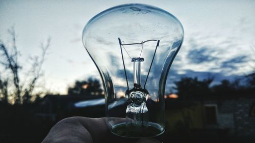 Close-up of human hand holding light bulb against sky during sunset