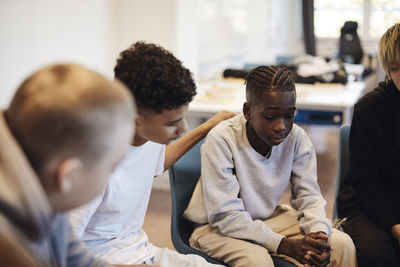 Sad teenage boy sitting amidst male friends in group therapy