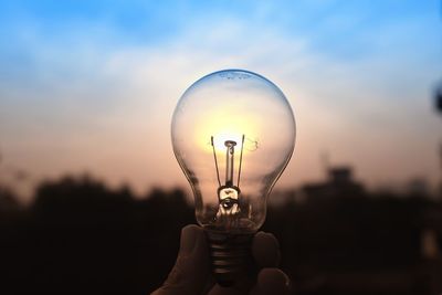 Cropped hand holding light bulb against sky during sunset