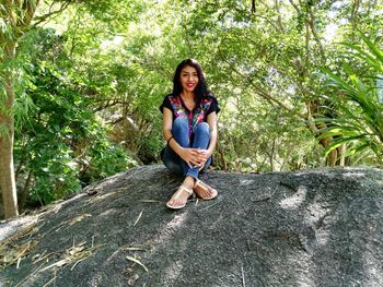 Portrait of smiling young woman sitting on rock against trees