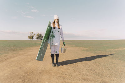 Young woman carrying green painting on dry field