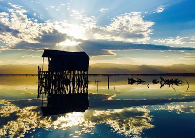 Silhouette stilt hut in lake against sunset sky