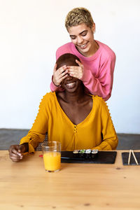 Smiling woman covering girlfriend eyes sitting at table