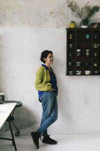 Smiling female owner with hand in pocket leaning on wall of workshop