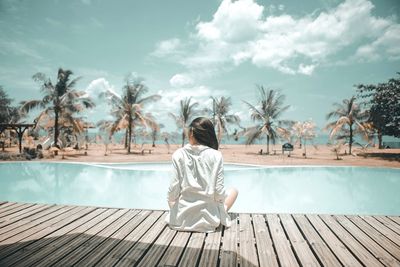 Rear view of woman sitting by swimming pool against sky