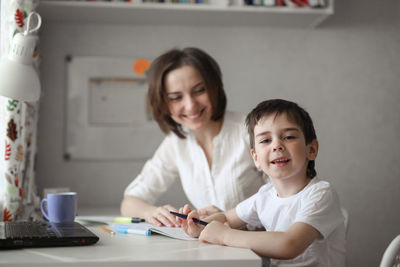 Beautiful woman tutor is engaged with a child boy at home at a table, real interior