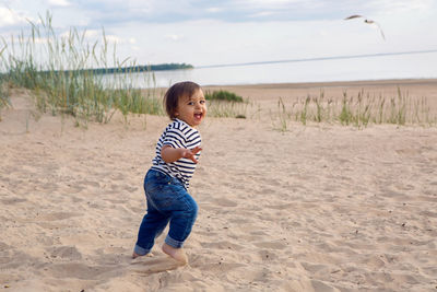 Baby boy in striped sailor t-shirt running on the sandy beach with seagulls near the sea in summer