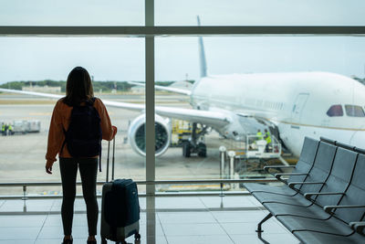 Woman with suitcase in hand, looks out the airport window while waiting for her flight.