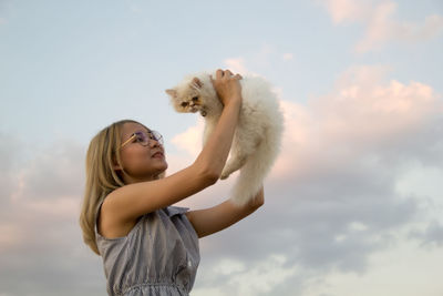 Young woman lifting cat against sky