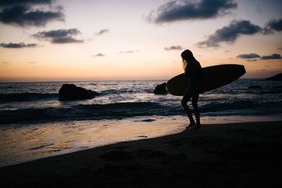 Silhouette woman with surfboard on beach against sky during sunset