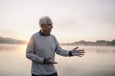 Senior man gesturing while standing against lake during sunrise