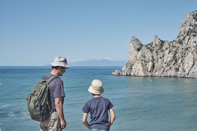 A boy and his grandfather standing on the shore of the bay, talking, looking at the rock. 