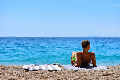 Rear view of shirtless man sitting by sea against clear sky