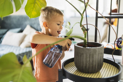 Cute boy sticking out tongue and watering potted plant at home