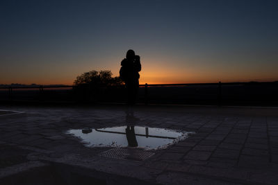 Silhouette of woman photographing while standing against sky during sunset