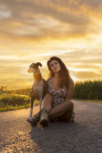 Portrait of woman with dog sitting on road against sky during sunset