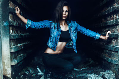 Portrait of confident young woman wearing denim jacket while crouching amidst walls