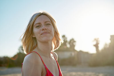 Positive blond female in swimwear looking at camera with smile while spending time on sunlit beach in weekend morning