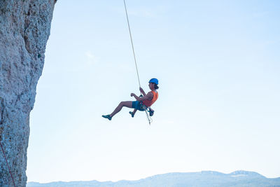 Low angle view of man handing on rope by rocky mountain against clear sky