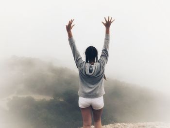 Rear view of teenage girl with arms raised standing on mountain during foggy weather