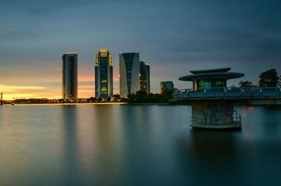 Illuminated buildings by river against sky during sunset