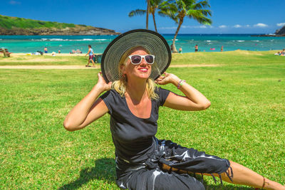 Portrait of happy woman wearing hat while sitting at grassy beach during sunny day