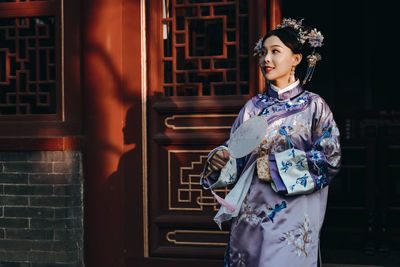 Thoughtful woman wearing kimono holding mirror while standing against built structure