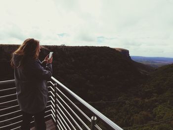 Side view of woman photographing landscape while standing on observation point against cloudy sky 