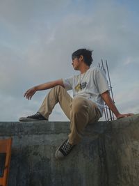 Young man sitting on rock against sky