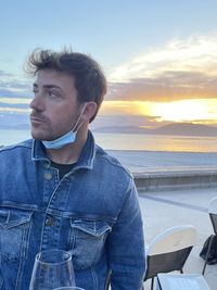 Man looking away while standing by sea against sky during sunset