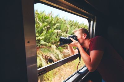 Man photographing through train window by trees