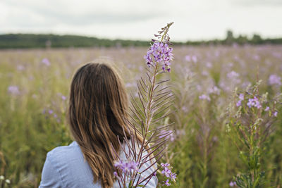 Young beautiful blond woman in purple shirt from behind in meadow among flowers of fireweed, nature
