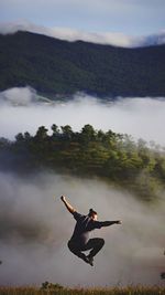 Rear view of man jumping against mountains during foggy weather