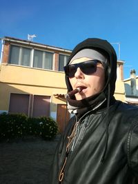 Low angle view of young man in hooded shirt smoking while standing by building against clear sky