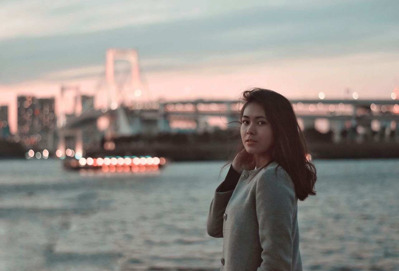 PORTRAIT OF BEAUTIFUL YOUNG WOMAN STANDING AGAINST SKY DURING SUNSET IN CITY