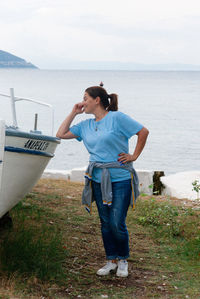 Full length of young woman with eyes closed and hand on hip standing by boat at shore against sky