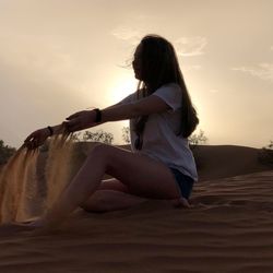 Side view of young woman sitting on land against sky during sunset