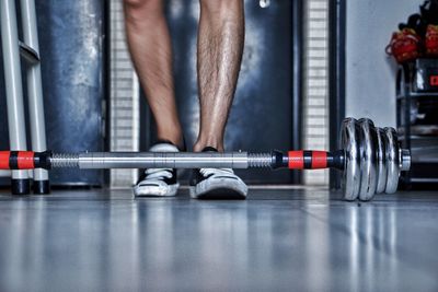 Low section of man standing by dumbbell on tiled floor in gym