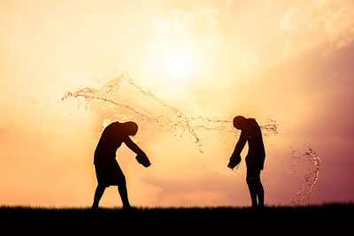 Silhouette friends splashing water with containers while standing on field against orange sky