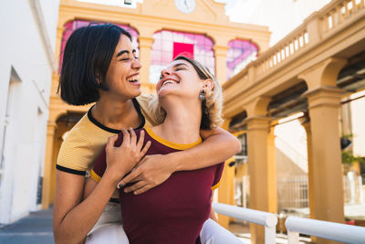 Smiling woman piggybacking girlfriend standing against building