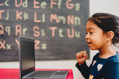 Side view of diverse pre school age girl at home doing distance learning on laptop computer