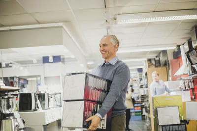 Smiling mature salesman with boxes walking in electronics store