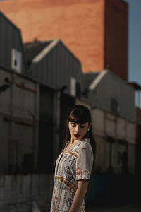 Side view of pensive young ethnic lady with long dark hair in casual dress standing in street near old houses and looking down on sunny day