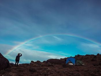 Rear view of woman with arms raised looking at rainbow while standing against sky