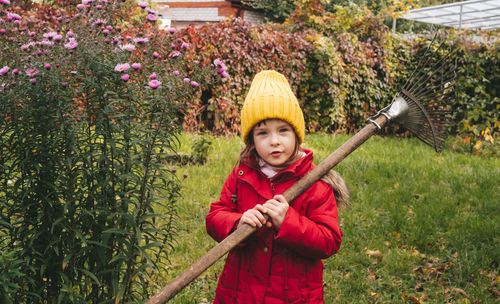 Girl in a red jacket and a yellow hat with a rake in the backyard. seasonal garden work. 