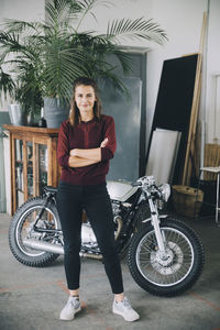 Portrait of confident female entrepreneur standing with arms crossed against motorcycle in creative office