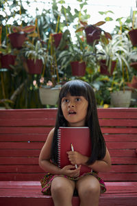 Cute girl holding book looking away against plants