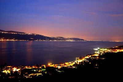 Illuminated city by sea against sky at night