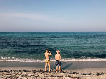 Rear view of shirtless boys standing at beach against sky