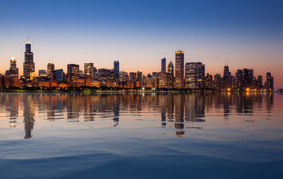 Illuminated buildings by lake in city against sky at dusk